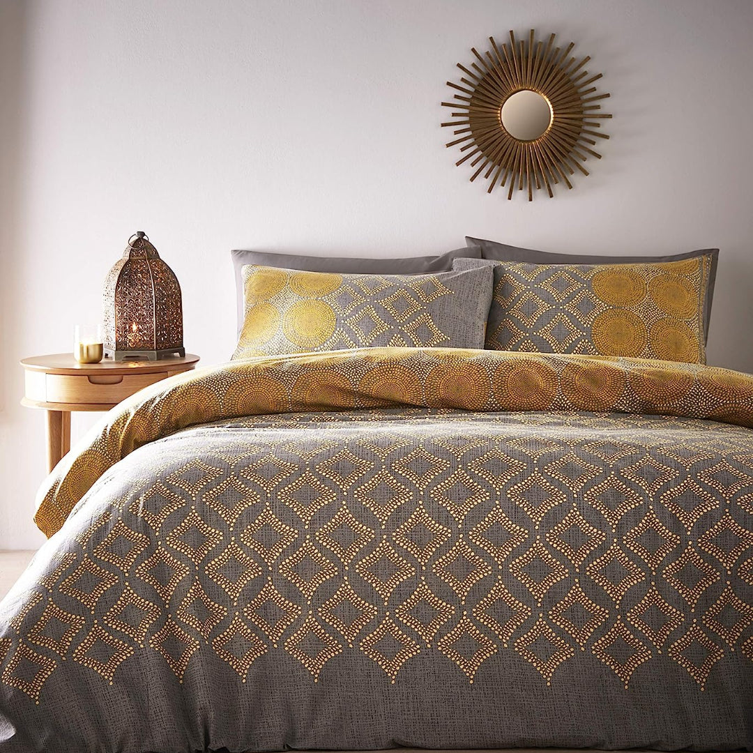 A styled bedroom scene showing a bed made up with a gray and mustard yellow patterned bedding set, including a duvet cover and matching pillows. A small wooden side table with a lantern and candle sits to the left, and a round sunburst wall mirror hangs on the wall above.