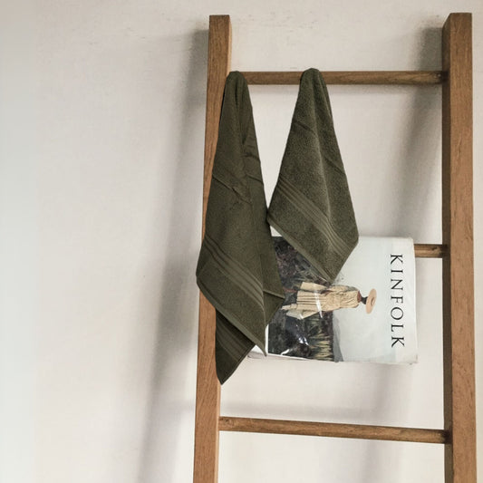 Wooden ladder with a sage green 500 gsm bath towel and a magazine against a plain wall.