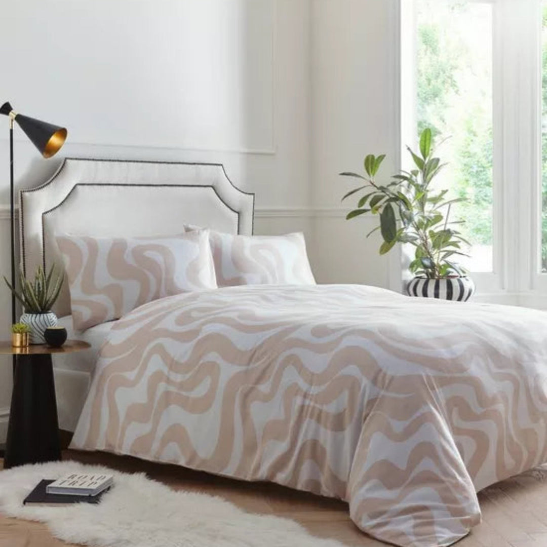 A bed dressed in a beige and white wavy patterned duvet cover and matching pillowcases, styled in a bright bedroom with a white headboard, black floor lamp, side table, and potted plant.