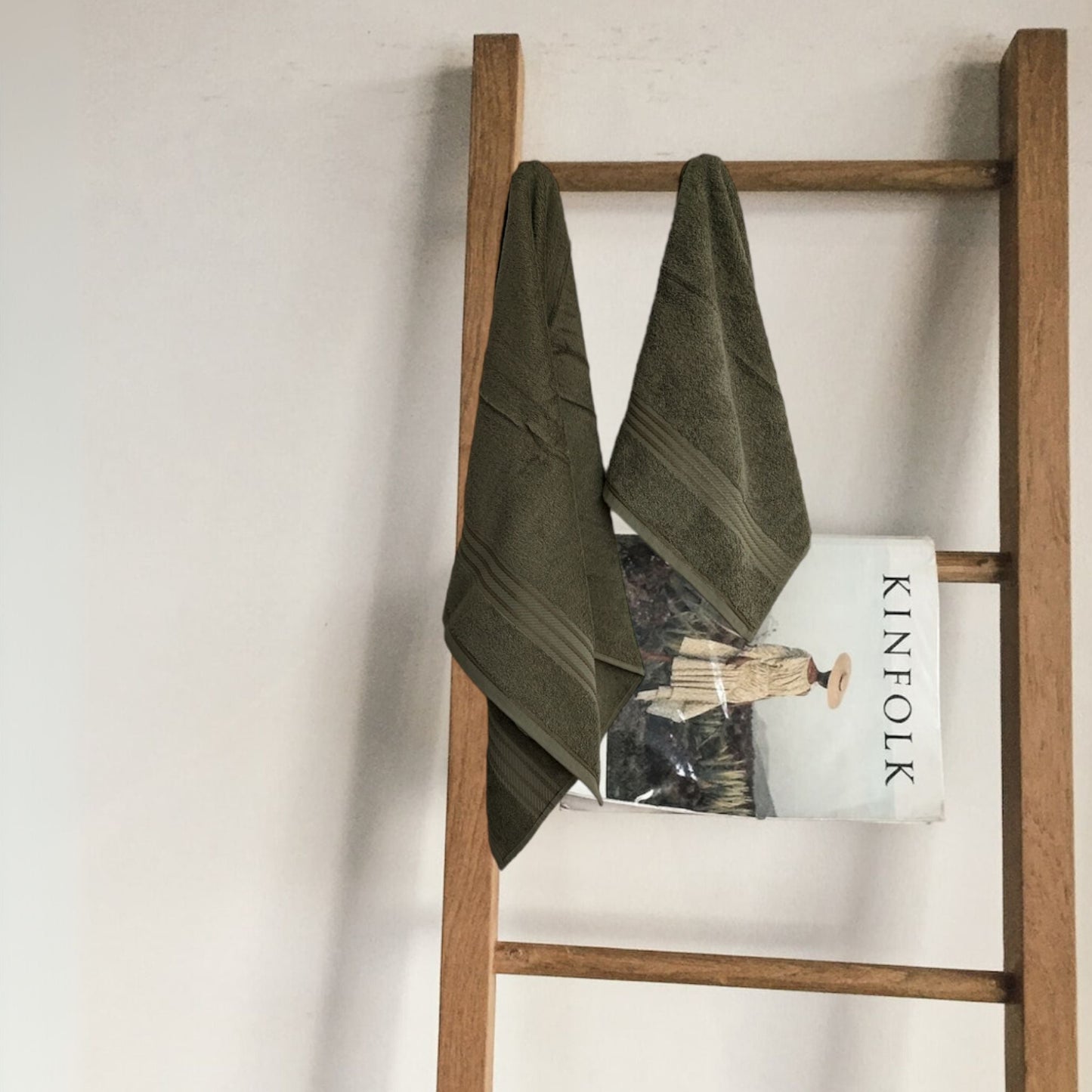 Wooden ladder with a sage green 500 gsm bath towel and a magazine against a plain wall.
