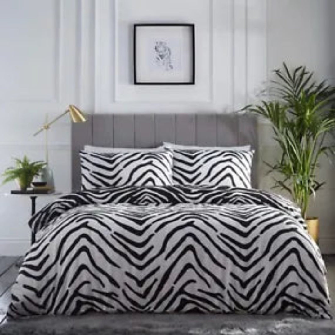 A bed dressed in a black and white zebra-patterned duvet cover and matching pillowcases, styled in a modern bedroom with a gray upholstered headboard, side table, lamp, framed wall art, and potted plants.